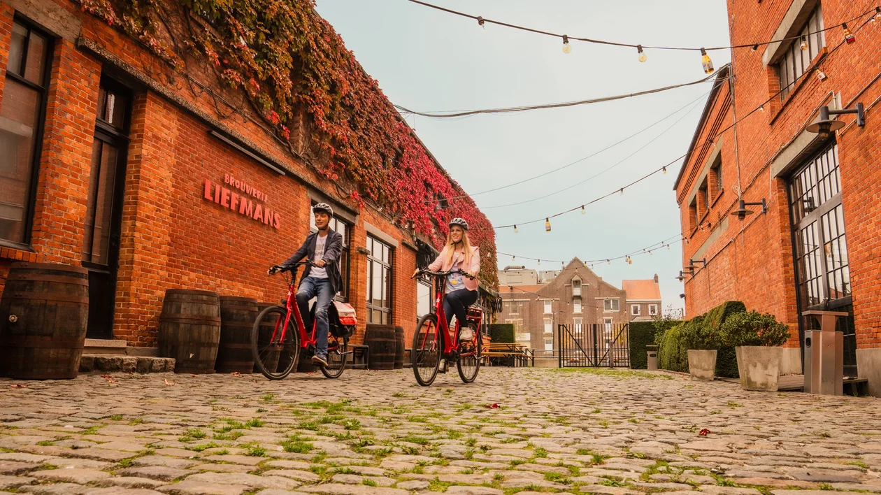 Two people riding bicycles down a cobblestone street lined with red brick buildings.