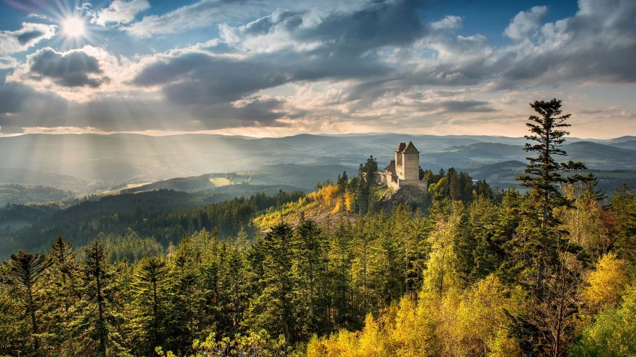 Castle on a hill surrounded by forest under a dramatic, sunlit sky.