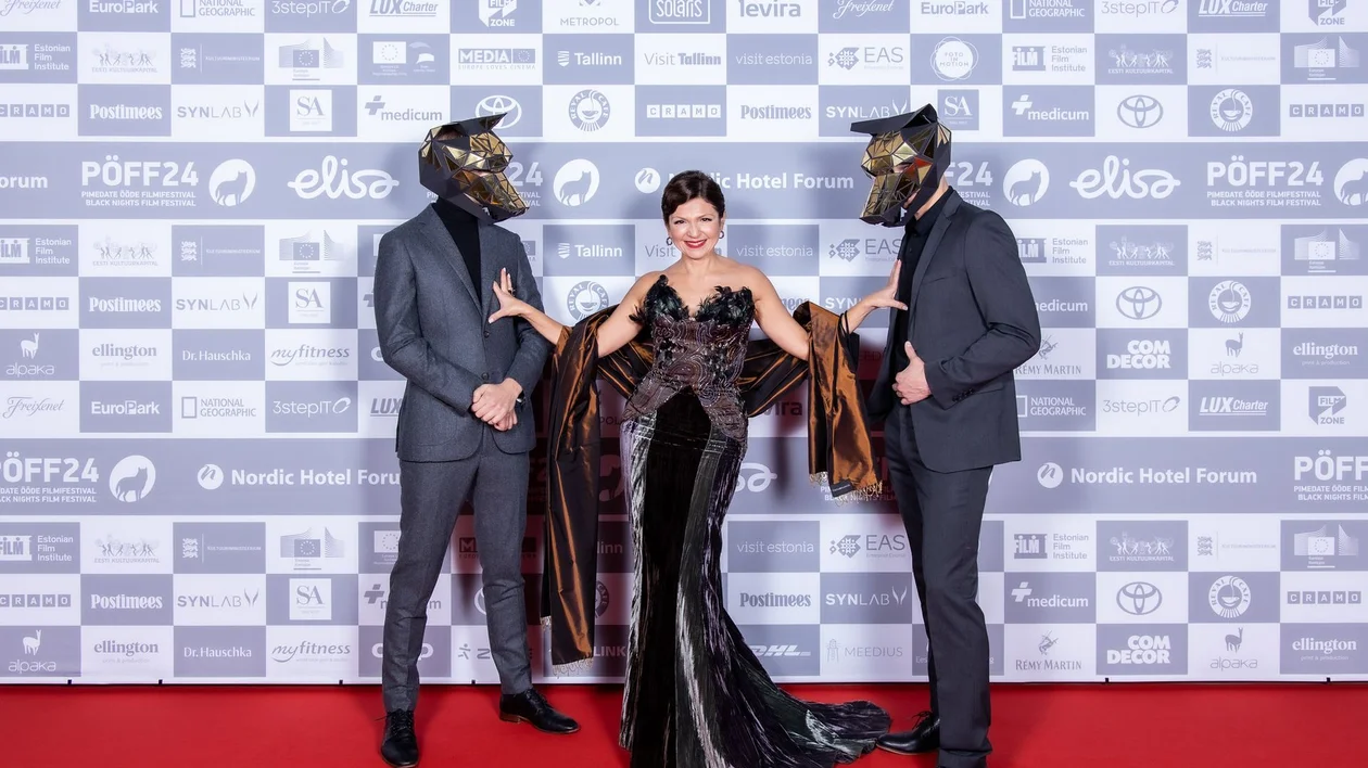 Three people in formal wear, two in wolf masks, posing on a red carpet.