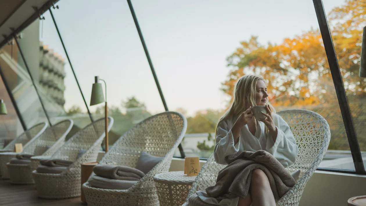 Woman in a robe sits in a wicker chair by large windows, drinking from a mug.