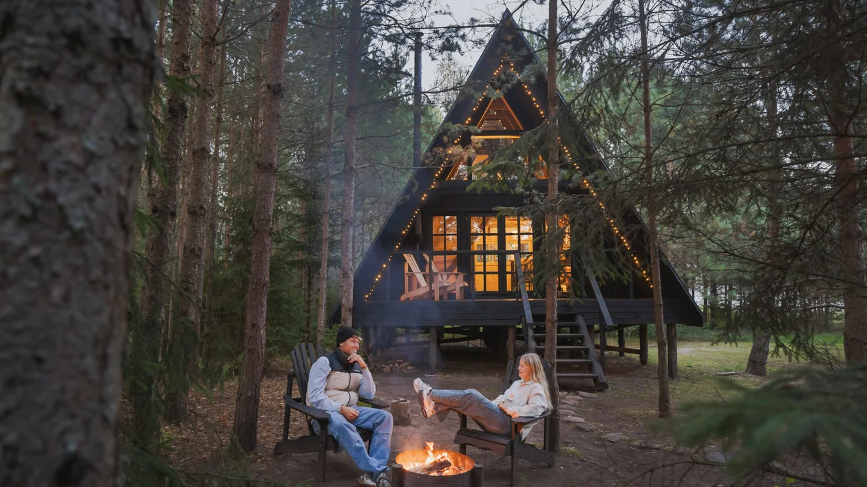 Two people relaxing by a campfire in front of a lit A-frame cabin in the woods.