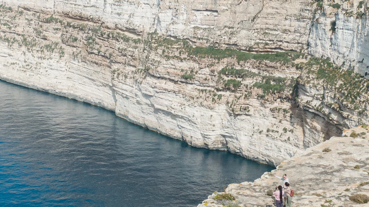 Steep coastal cliffs above blue sea with people walking on a rocky path.