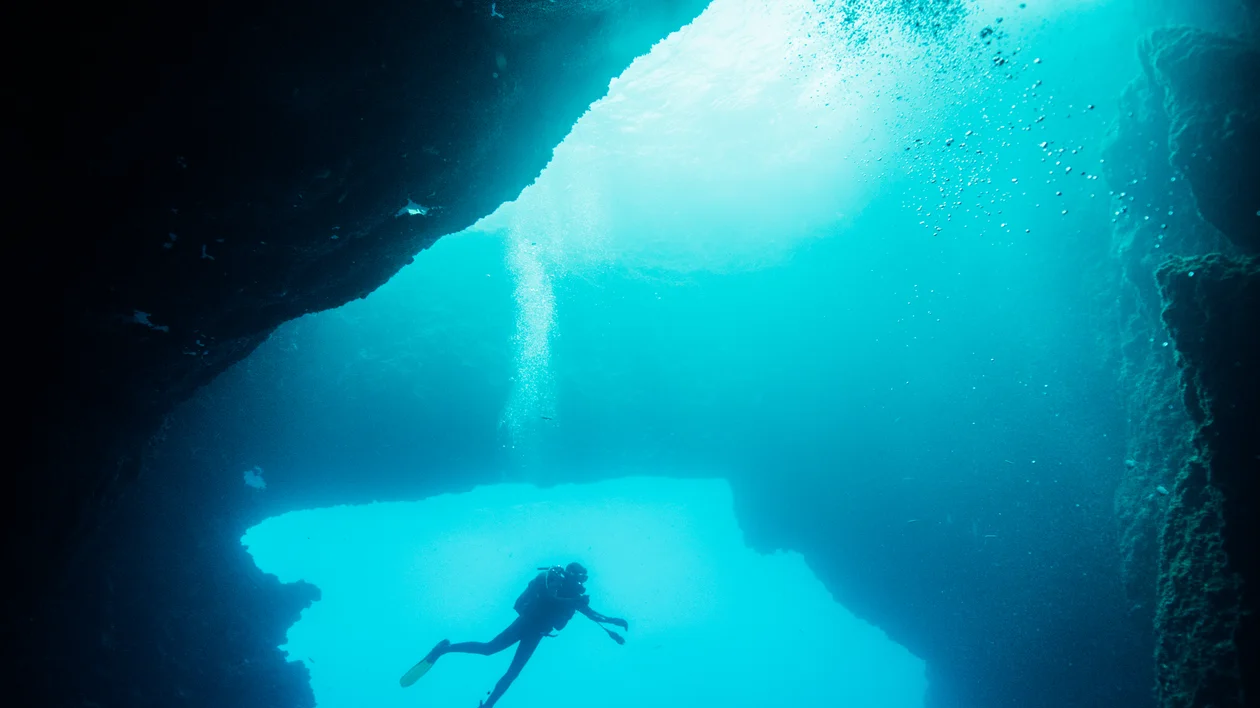 Scuba diver swimming inside an underwater cave with sunlight filtering through.