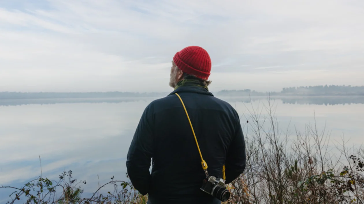 Person in a red beanie stands by a calm lake, camera hanging from their neck.