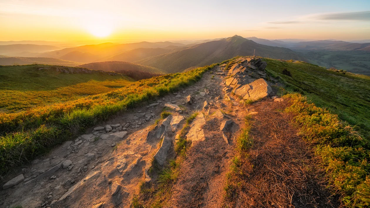 Rocky mountain trail at sunrise with green hills and distant peaks.