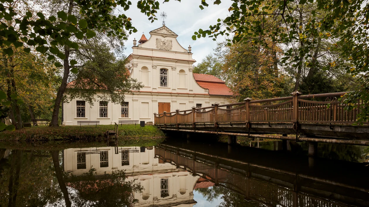 Historic building with red roof by a wooden bridge, reflected in a calm pond surrounded by trees.
