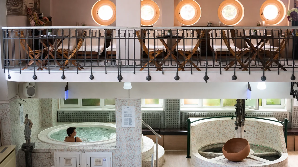Person relaxing in a spa jacuzzi below a balcony with empty tables and chairs.