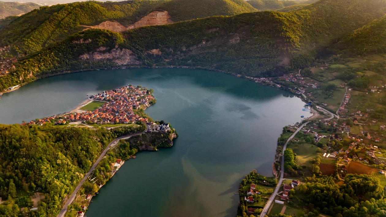 Aerial view of a lakeside village surrounded by green hills and winding roads.