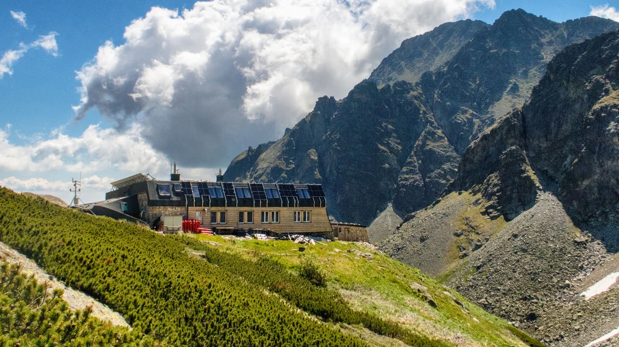 Mountain lodge with solar panels on a grassy slope, rugged peaks and clouds in the background.
