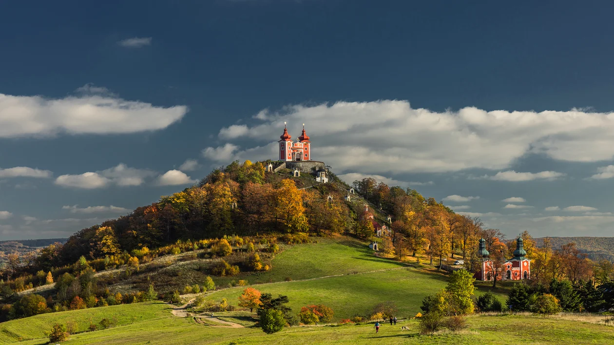 Hilltop church with red towers surrounded by autumn trees and green fields under blue sky.