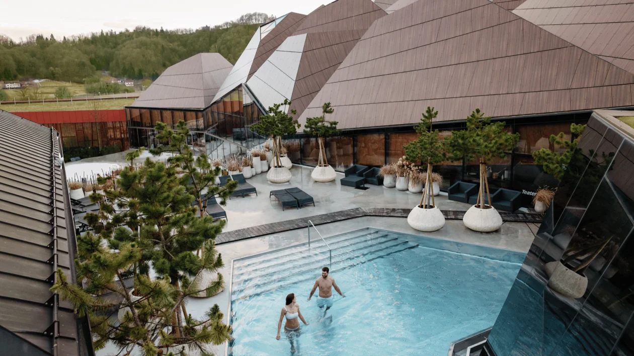 Two people in an outdoor pool surrounded by modern geometric buildings and greenery.
