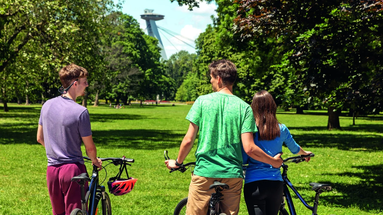 Three people with bikes in a sunny park, trees and a tower visible in the background.