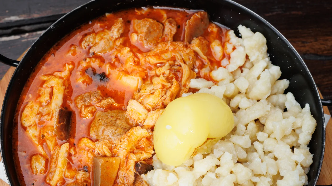 Stew with meat and red sauce served with dumplings and a poached egg in a black bowl.