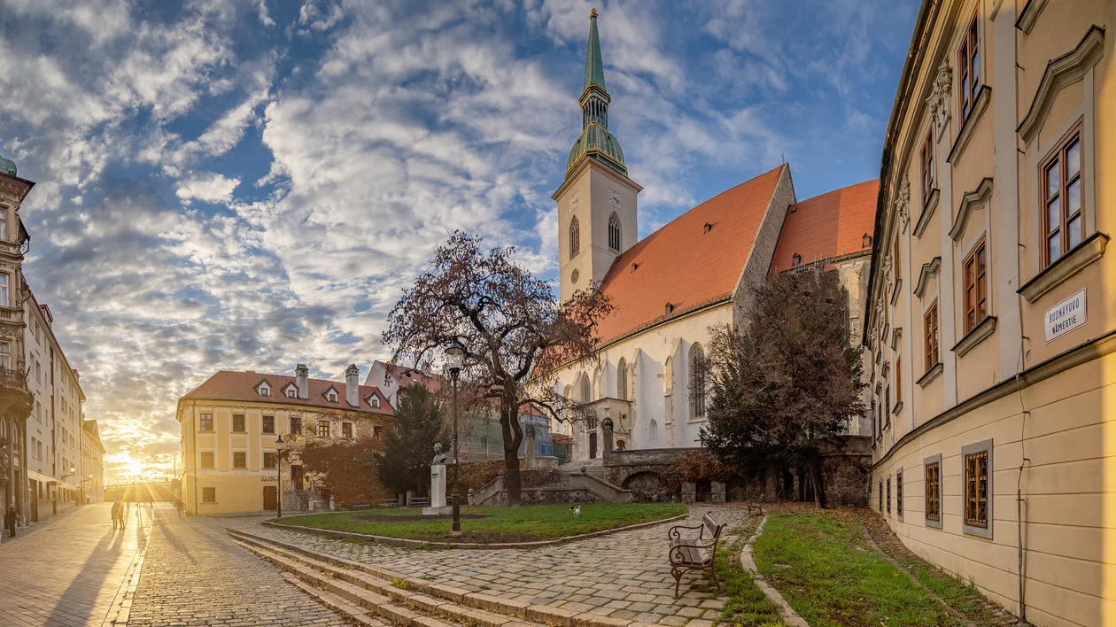 Historic church with tall spire and red roof at sunset, surrounded by old buildings and trees.