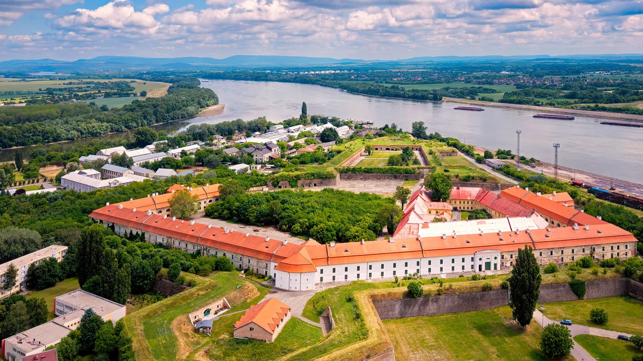 Large historic fortress with red roofs, surrounded by greenery, near a wide river under a blue sky.