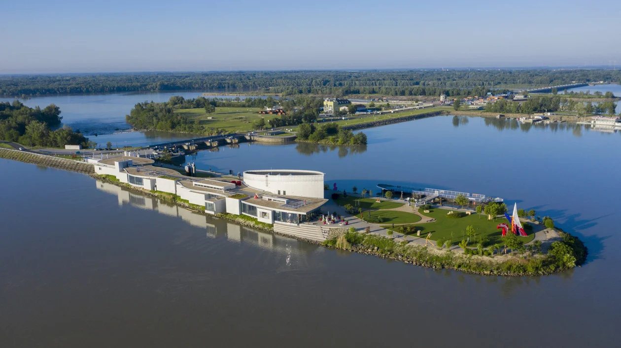 Modern riverside building complex with green lawns surrounded by water on a sunny day.