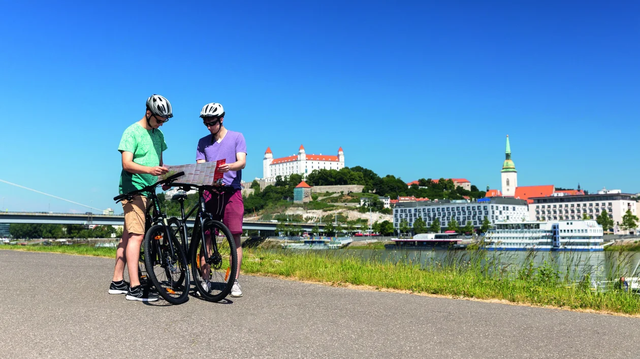Two cyclists with helmets look at a map by a river, cityscape and castle in background.