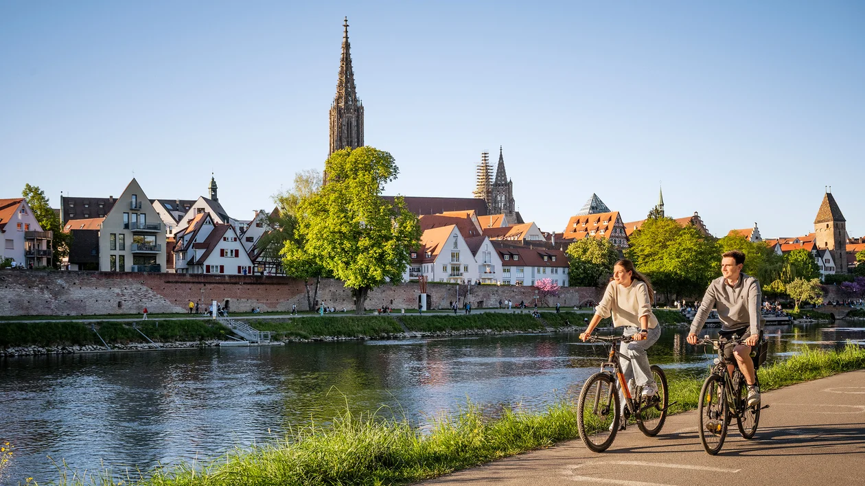 Two people cycling by a river with a cityscape and church in the background.