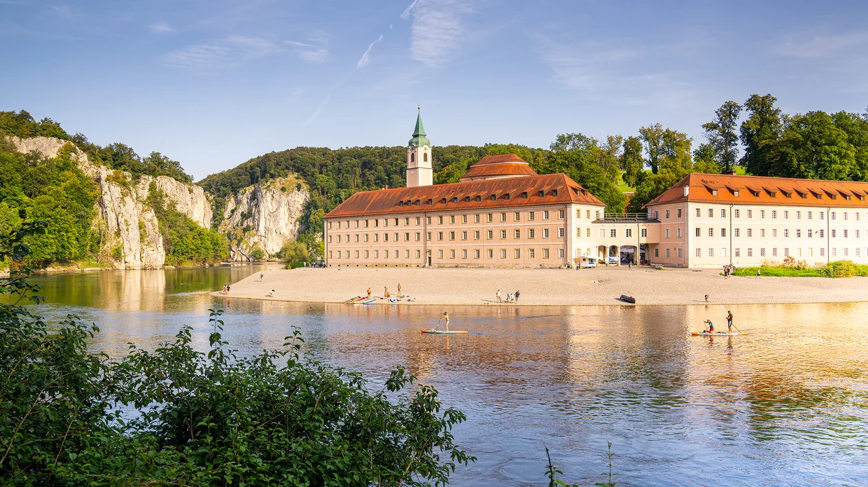 Large historic building with a tower beside a river, people walking on the sandy bank.