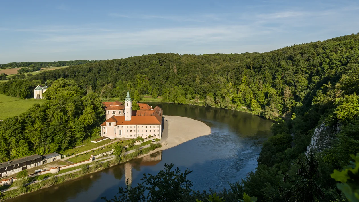 Monastery with red roof by a river bend, surrounded by dense green forest and hills.