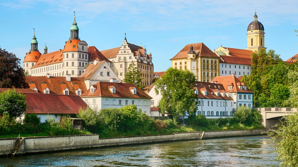 Historic European buildings with red roofs beside a river, trees in the foreground.