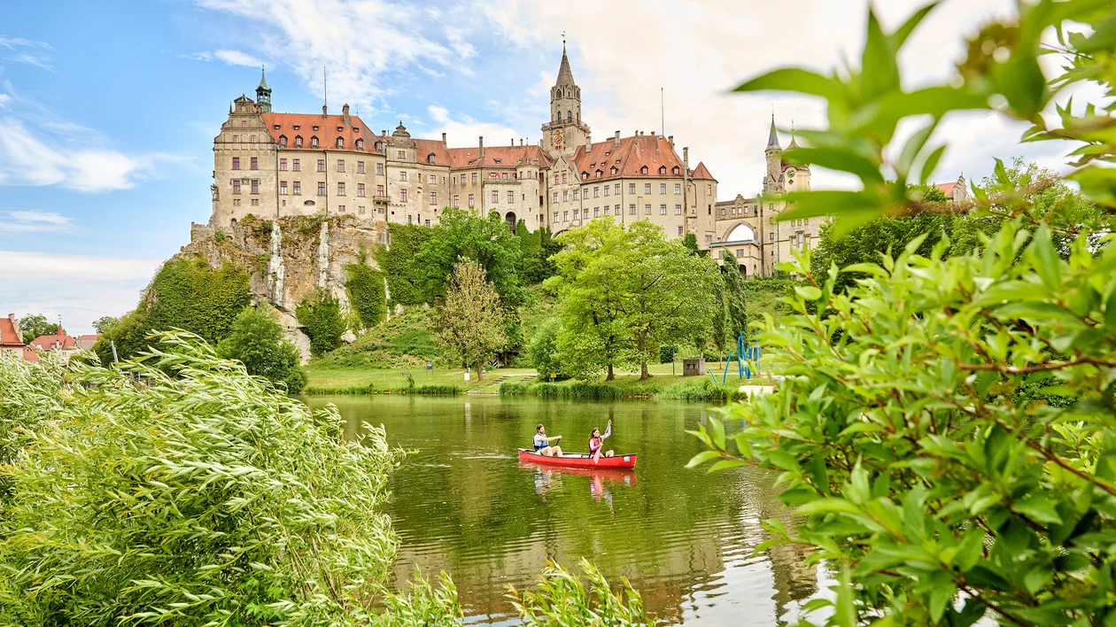 Large castle on a hill, trees surrounding, two people canoeing on a calm river in front.