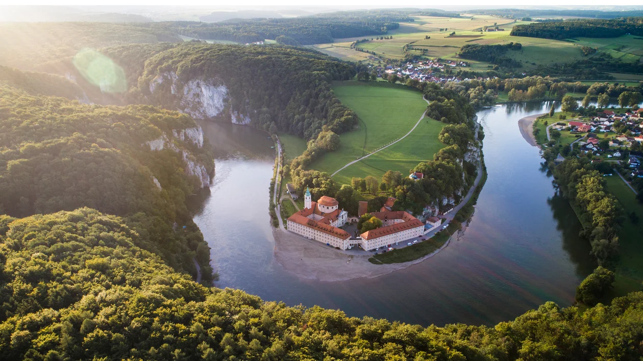 Aerial view of a river bending around a monastery surrounded by forests and fields.