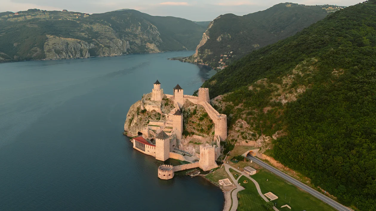 Medieval fortress on a riverbank surrounded by green hills and calm water.