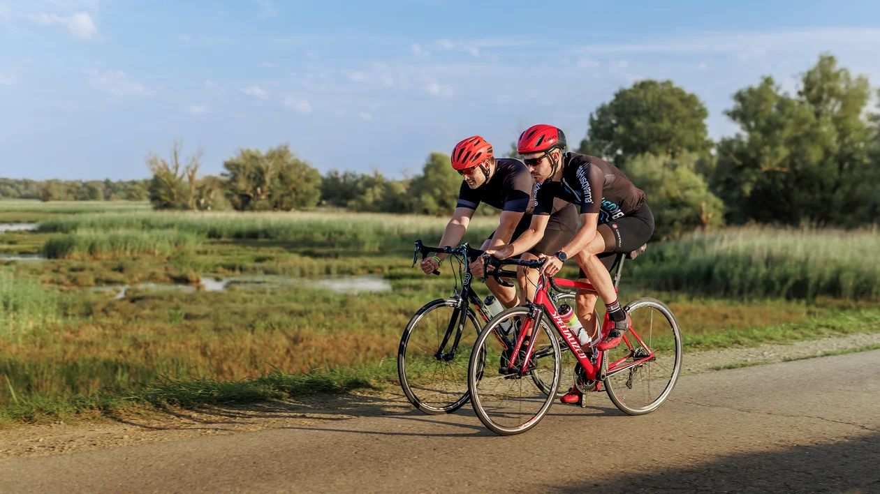 Two cyclists in red helmets ride bikes on a rural path beside green fields and wetlands.