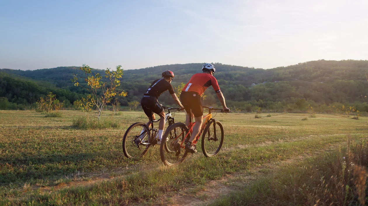 Two cyclists ride on a dirt path through a sunny, grassy landscape with hills in the background.