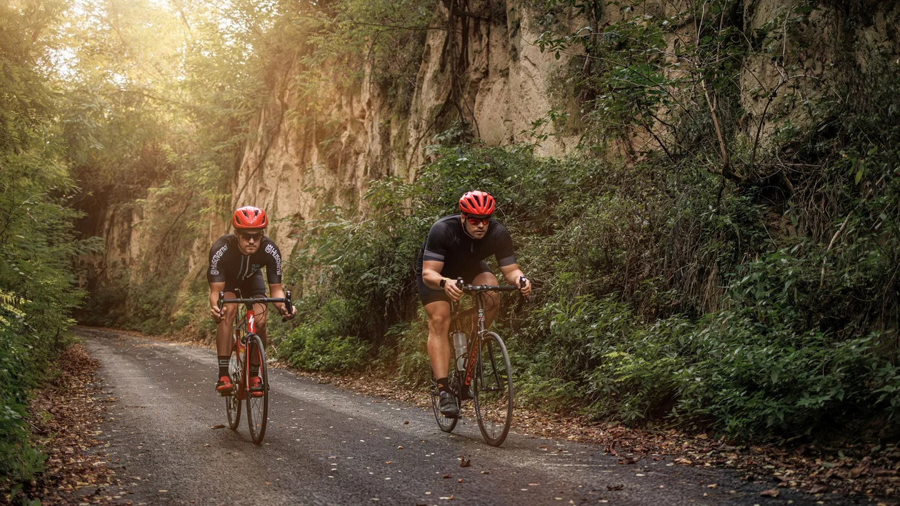 Two cyclists riding on a forest path beside rocky cliffs in warm sunlight.