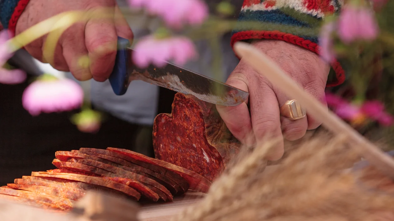 Hands slicing cured sausage with a knife on a cutting board, flowers in the background.