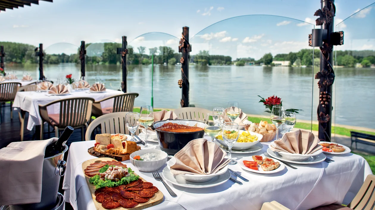 Outdoor restaurant table set with food by a river, surrounded by trees and clear sky.