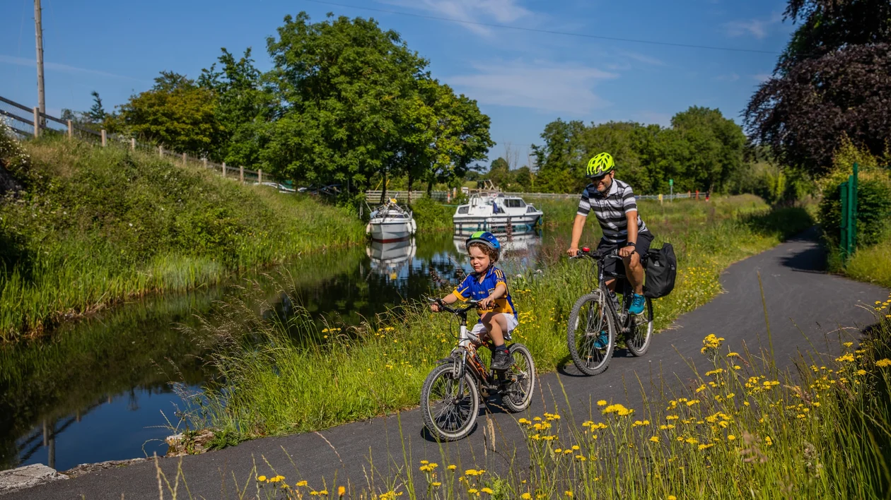 Adult and child cycling on a sunny path beside a canal with boats and wildflowers.