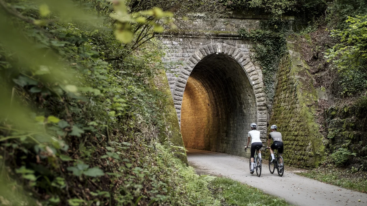 Two cyclists entering a stone tunnel surrounded by greenery.
