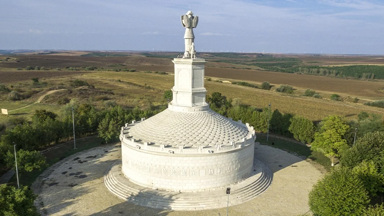 Large white circular monument with a statue on top, surrounded by trees and open fields.