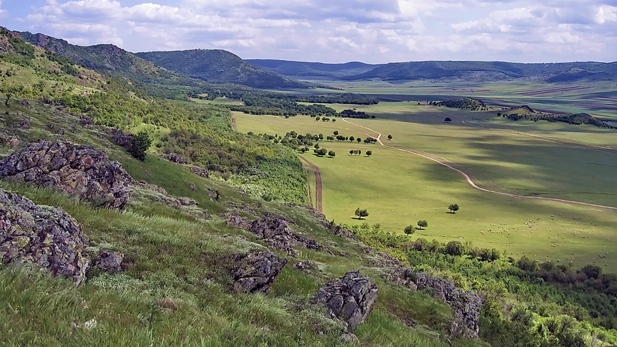 Rolling green hills and rocky slopes overlook a vast sunlit valley under a blue sky with clouds.