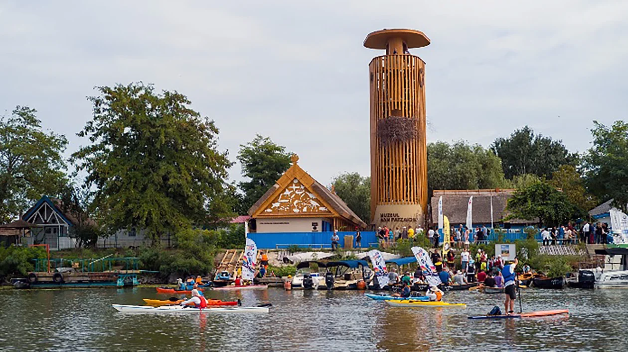 People kayaking on a river near a tall wooden tower and buildings with trees in the background.