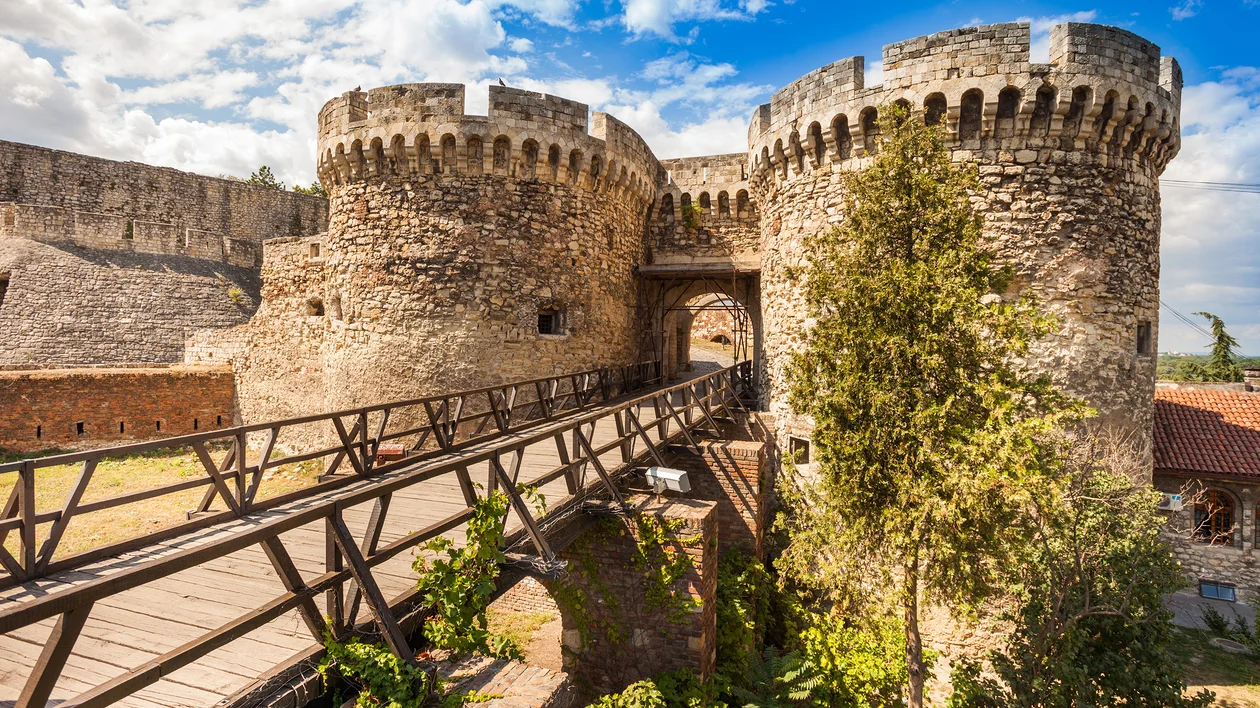 Stone fortress with round towers, wooden bridge, and partly cloudy sky in the background.