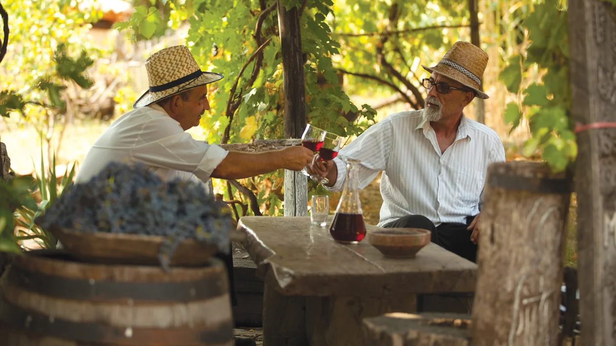 Two men in straw hats clinking wine glasses at a rustic outdoor table.