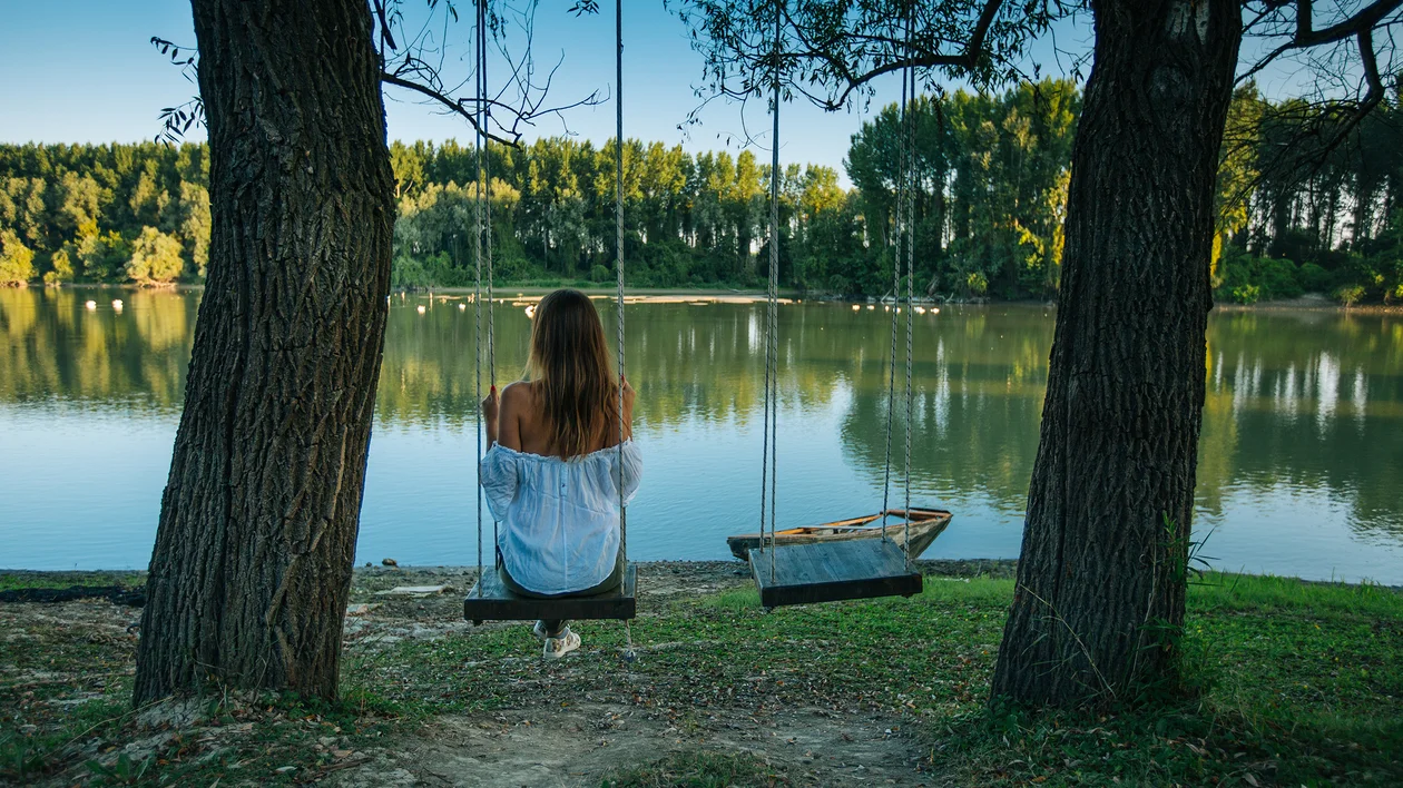 Woman sitting on a swing by a lake, surrounded by trees, facing the water.