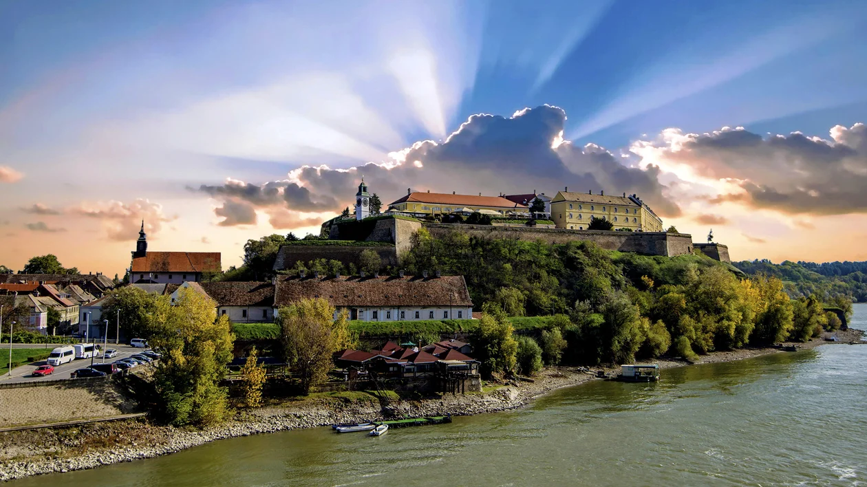 Hilltop fortress with sun rays breaking through clouds, overlooking a river and town.