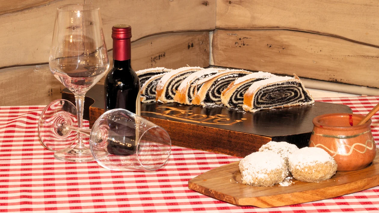 Sliced poppy seed roll, red wine, cookies, and two glasses on a checkered tablecloth.