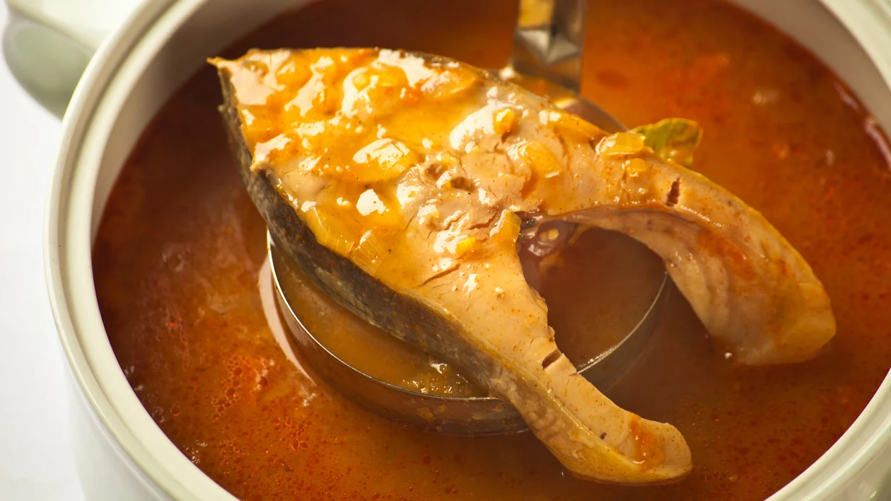 Fish steak in reddish soup in a white bowl, served with a metal ladle.