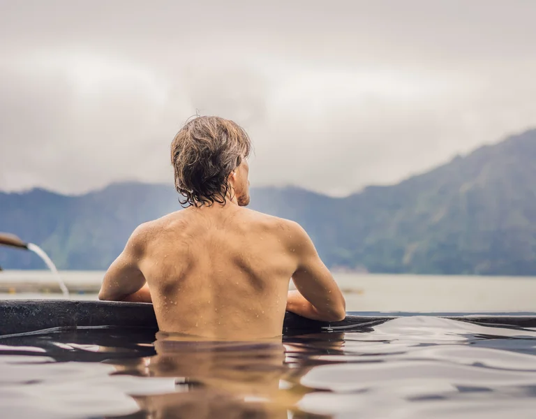 Man relaxing in a hot spring with mountains and cloudy sky in the background.