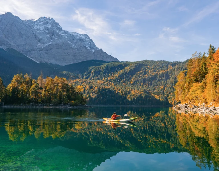 Canoe on a clear lake surrounded by autumn trees and distant snow-capped mountains.