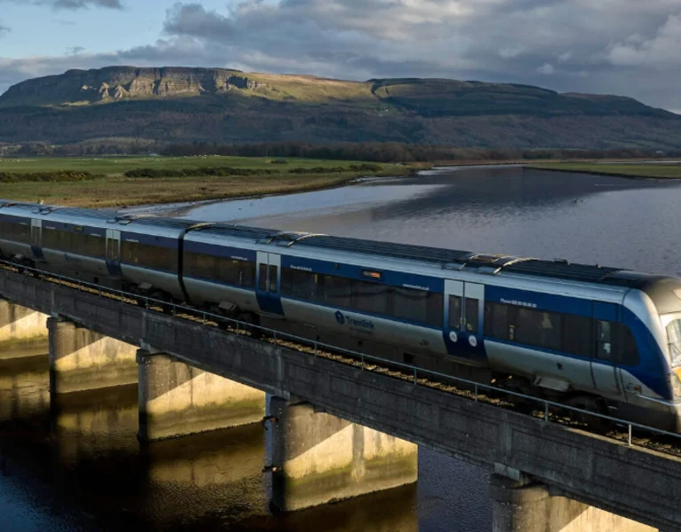 A train crosses a raised bridge over a river with a typical Irish landscape.