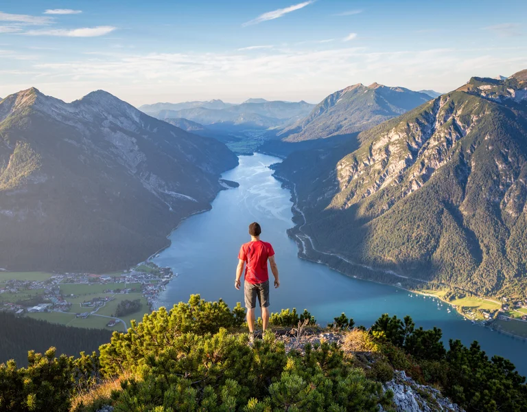 Person in red shirt stands on hilltop overlooking a lake between mountains at sunset.