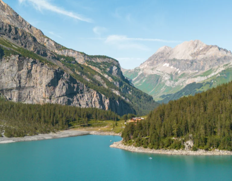 Scenic view of Oeschinensee in Switzerland, featuring turquoise waters surrounded by lush green forests and dramatic rocky mountains under a clear blue sky.