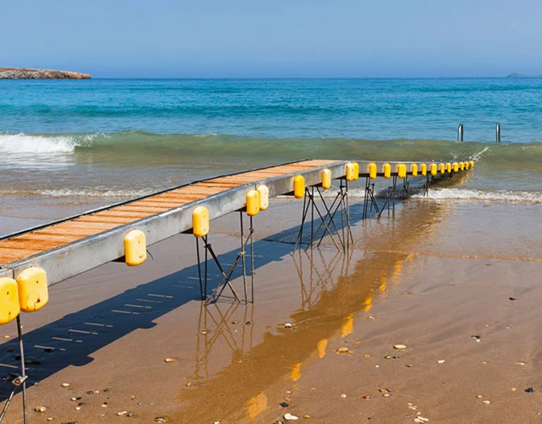 A floating dock extends into the sea on a sandy beach under a clear blue sky.